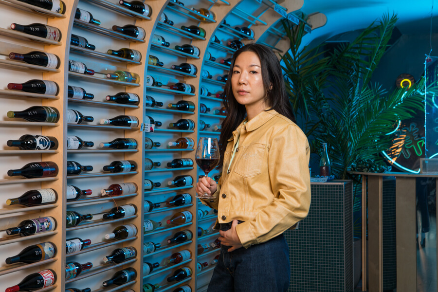 A photo of Grape Crush co-owner Nancy Chen beside a wall of bottles