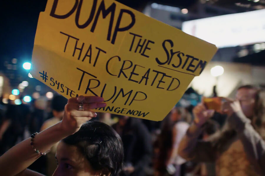 A placard reads Dump the system that created Trump" in a scene from The New Corporation