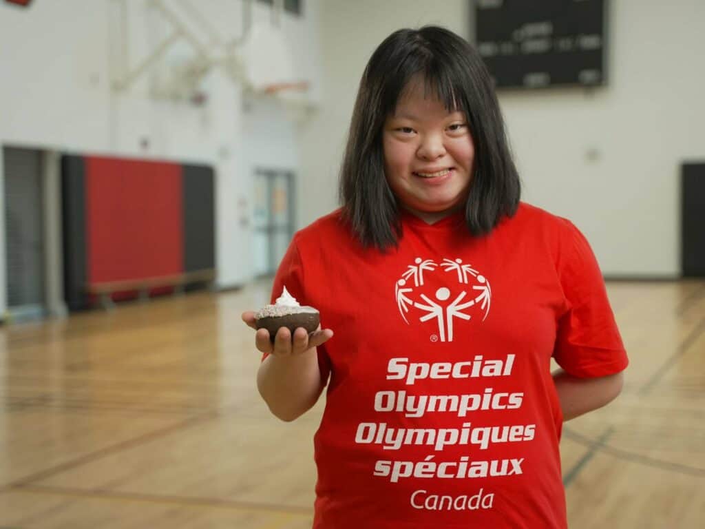 Joyful young girl holding a cupcake in a gymnasium during Special Olympics event in Canada.