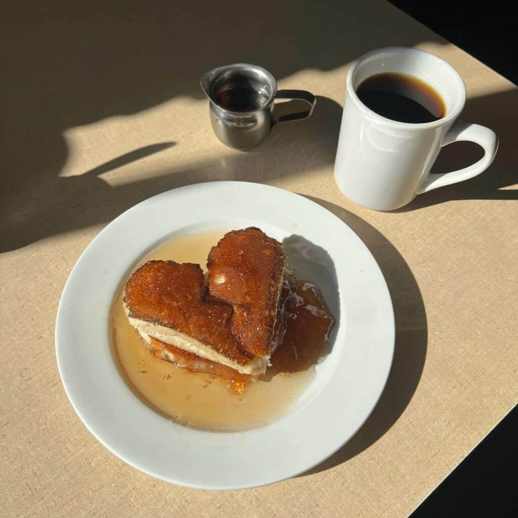 Rich, golden-brown French toast with caramel syrup served on a white plate, paired with a cup of black coffee and a small pitcher of syrup, under natural morning light.