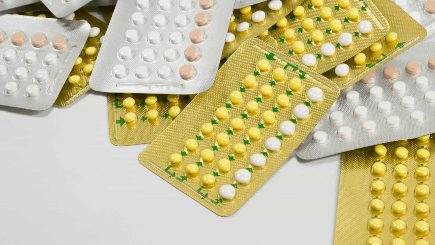 Blister packs of mixed pills and tablets, including yellow and white medications, stacked and scattered on a white surface, representing pharmaceuticals and medication supplies.