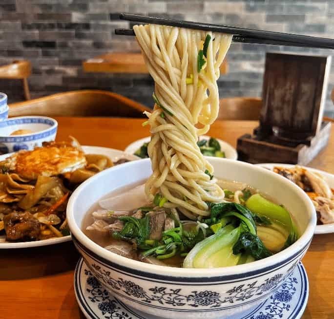Fresh steaming bowl of Asian noodle soup with green vegetables and herbs in a traditional ceramic bowl.