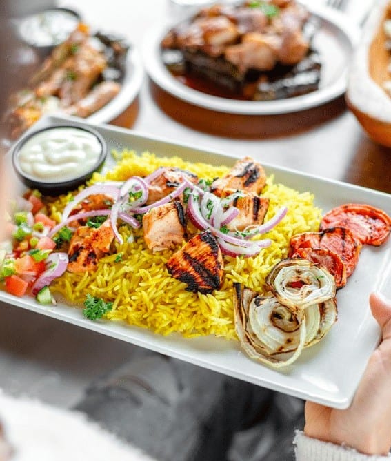 Colourful grilled chicken skewers and yellow rice on a white platter with fresh vegetables and dips, showcasing delicious Middle Eastern cuisine in Toronto.