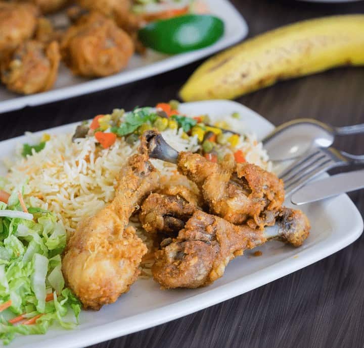 Crispy fried chicken wings served with rice and vegetables on a white plate, featuring a side salad and banana in the background.