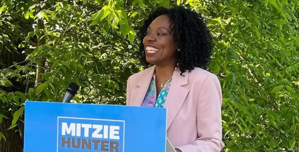 African American woman smiling at outdoor event, delivering speech at podium, lush green foliage background, political campaign, community engagement in Toronto.