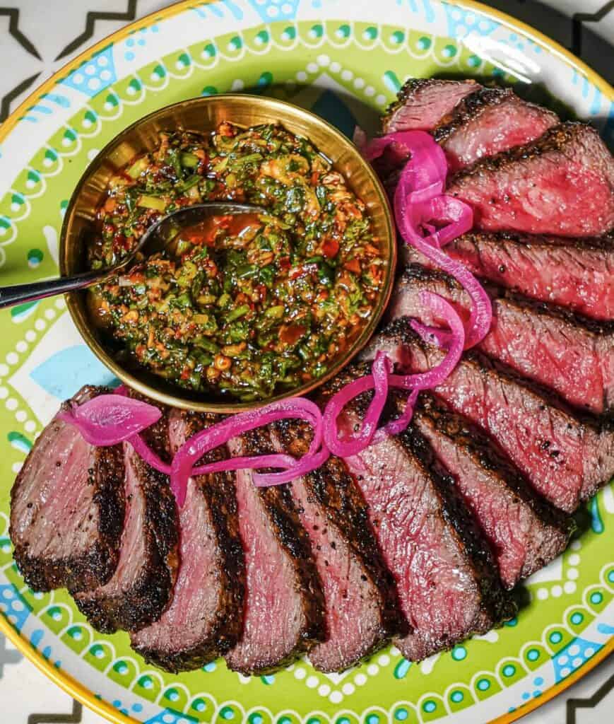 Thinly sliced grilled steak with chimichurri sauce and pickled red onions on a colorful plate, showcasing delicious Toronto barbecue cuisine.