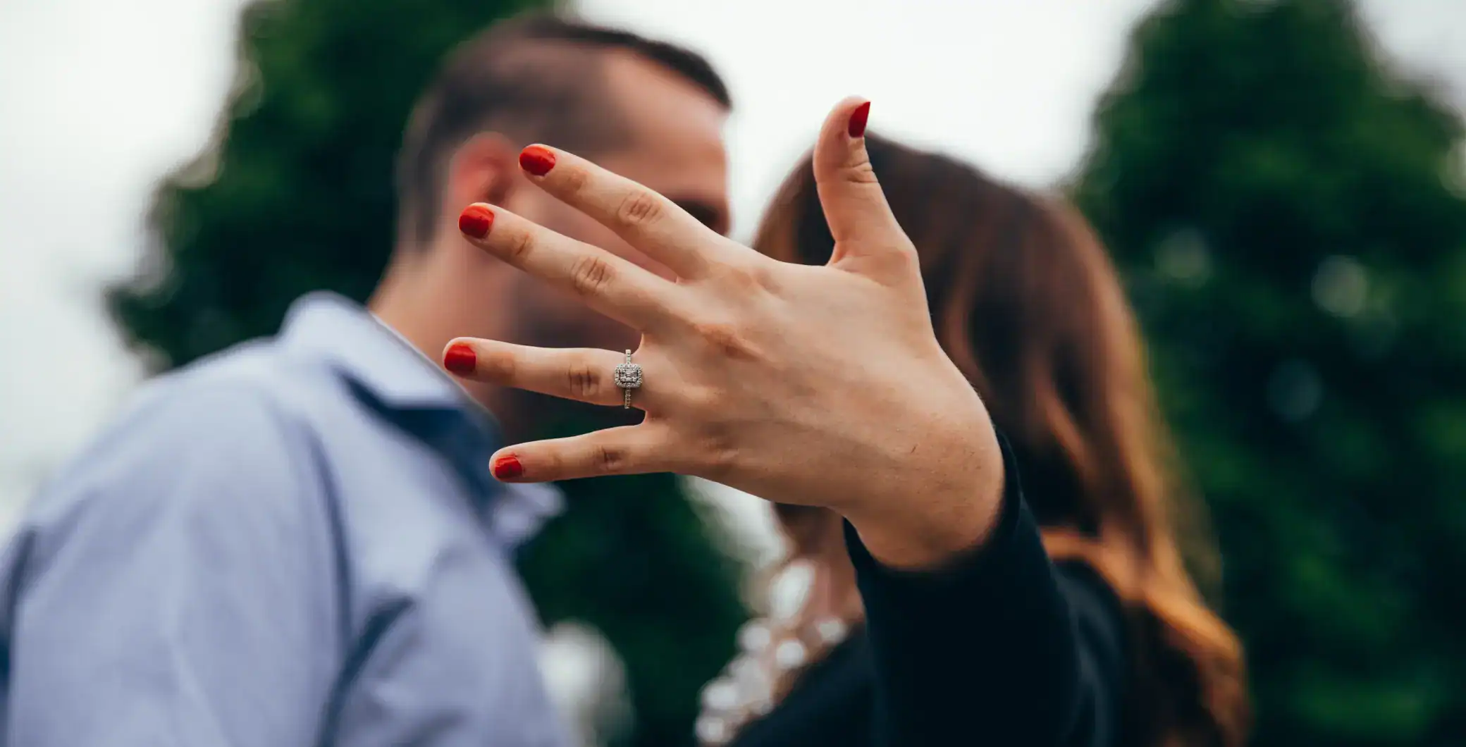 Close-up of a woman's hand showing an engagement ring with red painted nails, blurred couple in the background, outdoor setting, love, engagement, couple, Toronto.