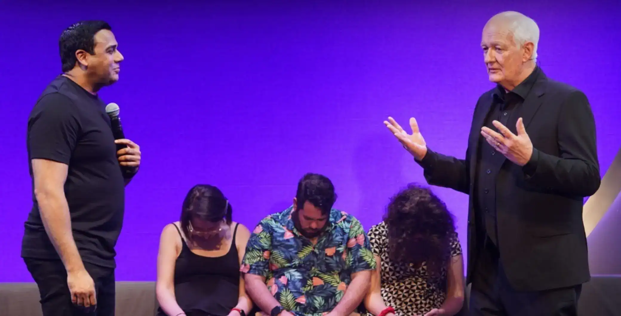 Vibrant indoor discussion with diverse group, including a man speaking into a microphone and a senior man gesturing, under purple lighting, at a Toronto event.