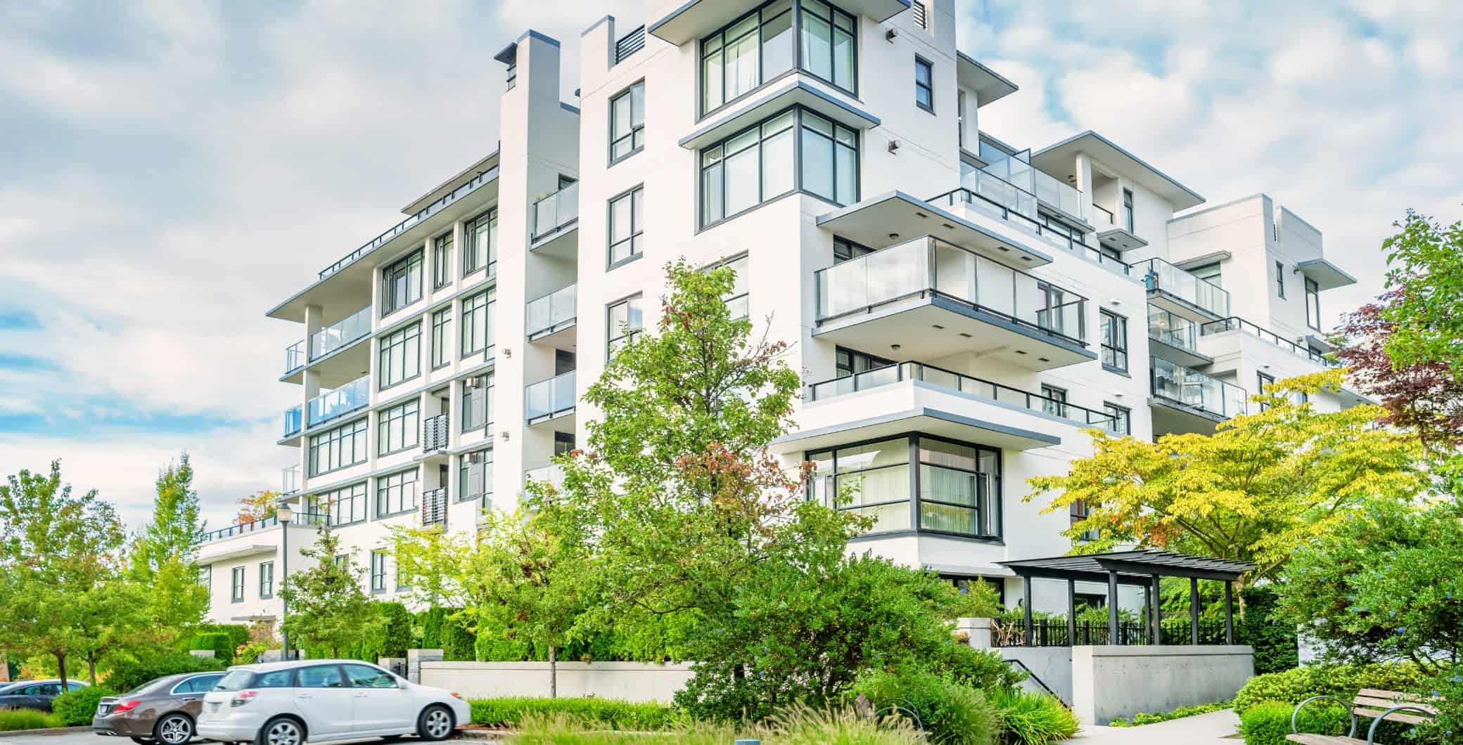 Modern white condominium building with large glass windows and balconies, surrounded by lush green trees and a parking lot in Toronto, Canada.