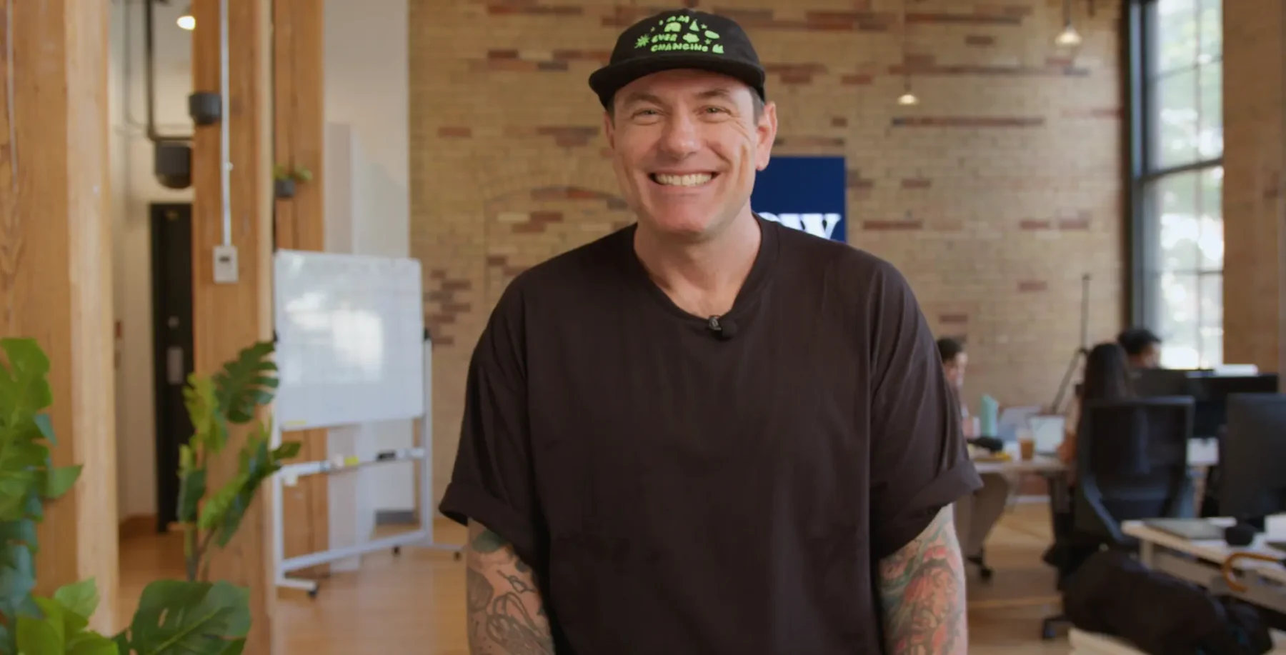 Bright, smiling man in a black t-shirt and cap with tattoos on his arms at NOW Toronto office during daytime.