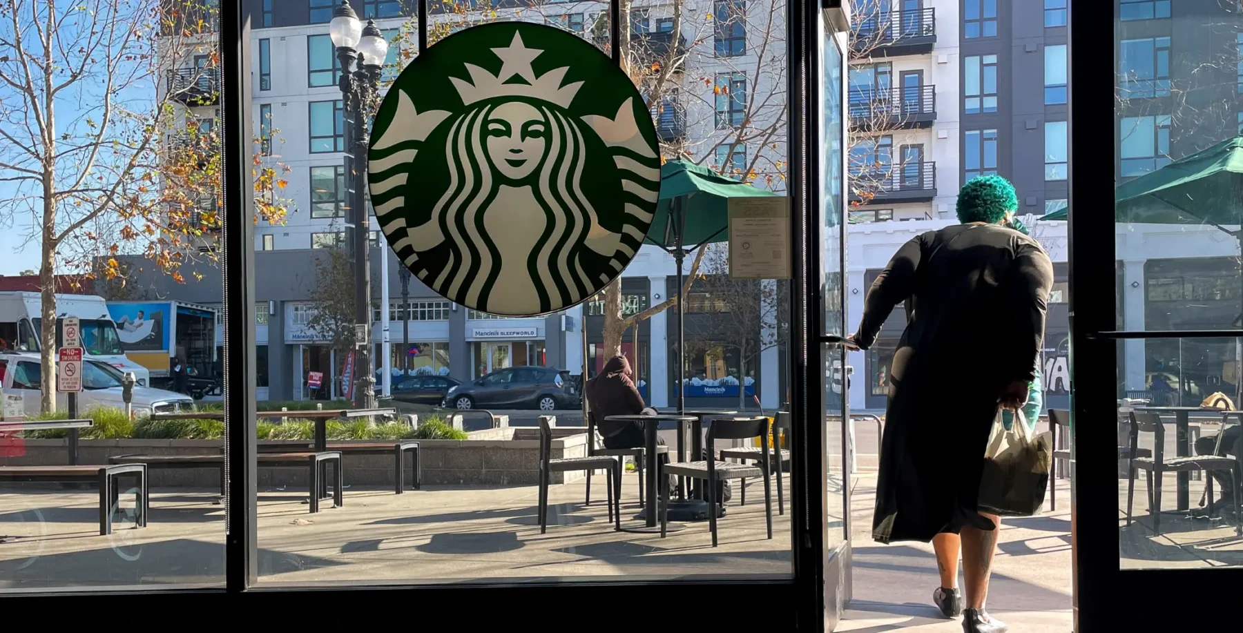 Coffee shop entrance with Starbucks logo, city street view, outdoor seating, and a person with turquoise hair entering the cafe.