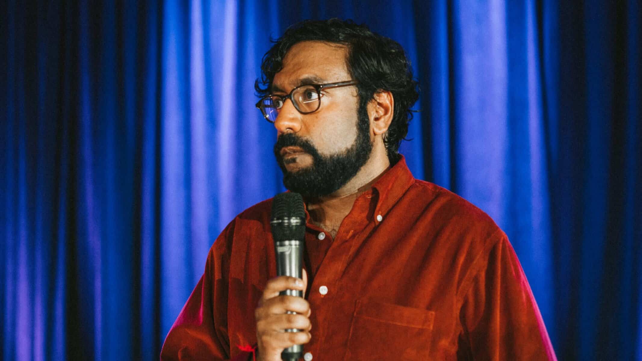 Man performing stand-up comedy on stage with blue curtains in Toronto, comedy event in Toronto, live entertainment in Toronto, NOW Toronto event.