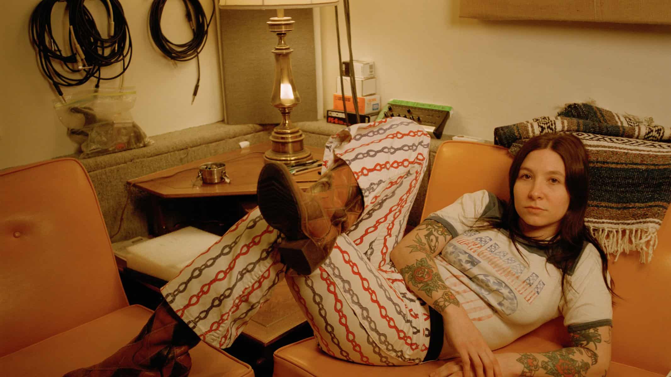 Discarded cables hanging on wall in a cluttered room with a young woman lying on chair, tattoos visible on arms, vintage decor and warm lighting, casual and artistic atmosphere.