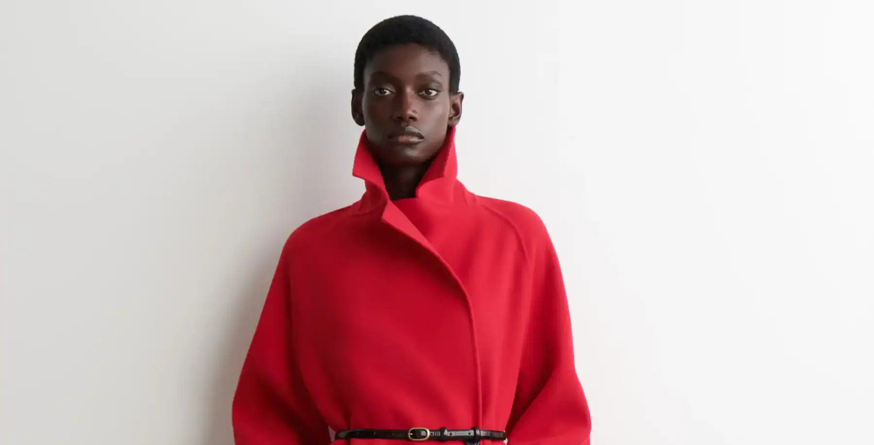 Modern black woman in a bold red coat, standing against a white wall, showcasing contemporary fashion and urban style in Toronto.