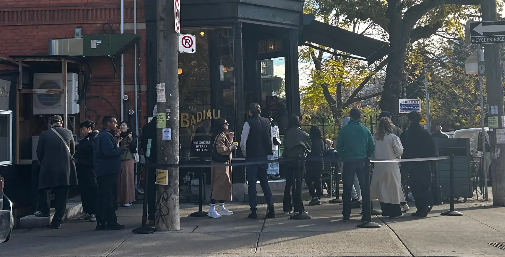 People queue outside Badali Toronto restaurant, enjoying takeout and outdoor dining on a busy city street corner.