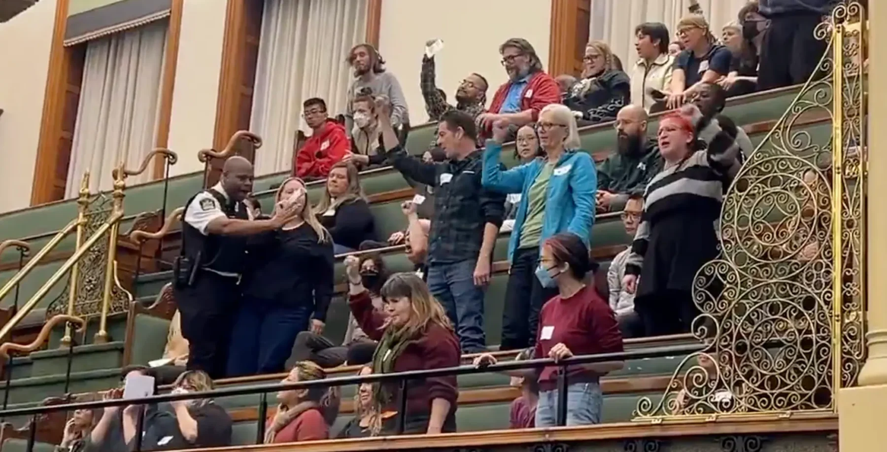 Diverse group of people raising hands during a public hearing or discussion in a historic Toronto theatre or auditorium setting.