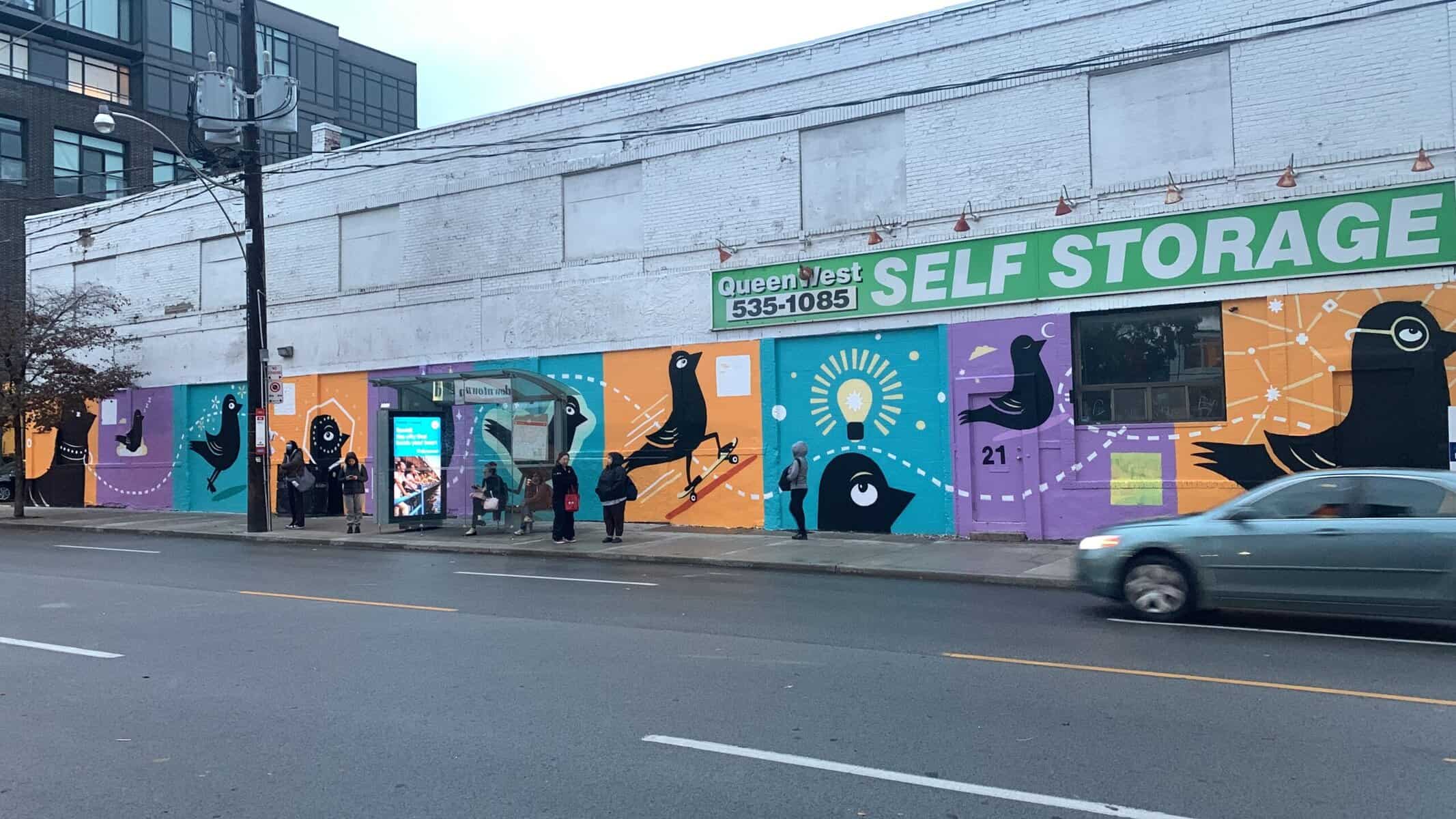 Vibrant colourful mural on exterior of a city building with bird illustrations and people waiting at a bus stop on Toronto street, showcasing urban art and community life in Toronto.