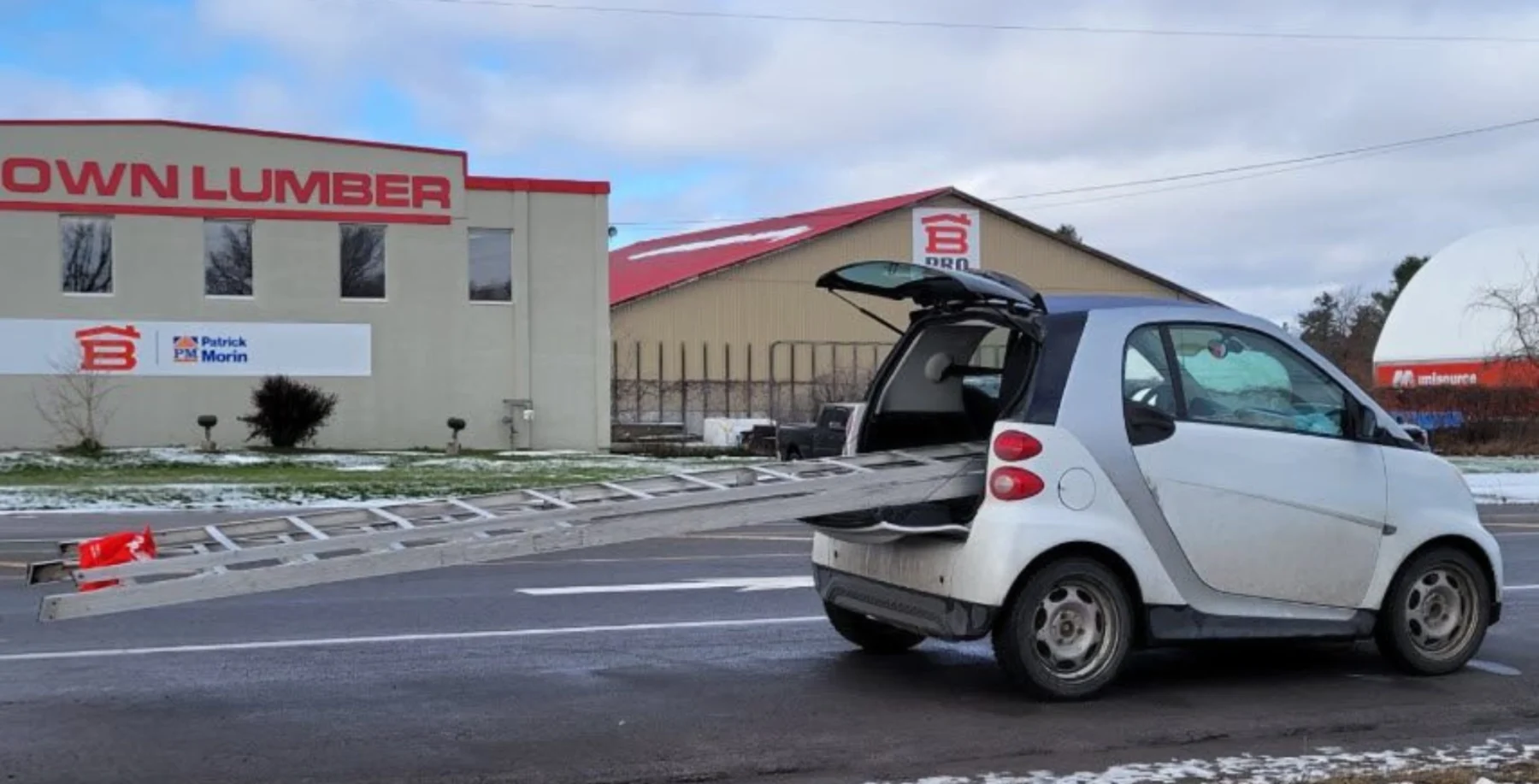 Small electric car with its trunk open, transporting a ladder on the road near a building with "DOWNLUMBER" signage in Toronto, winter season, overcast sky, snow on the ground.