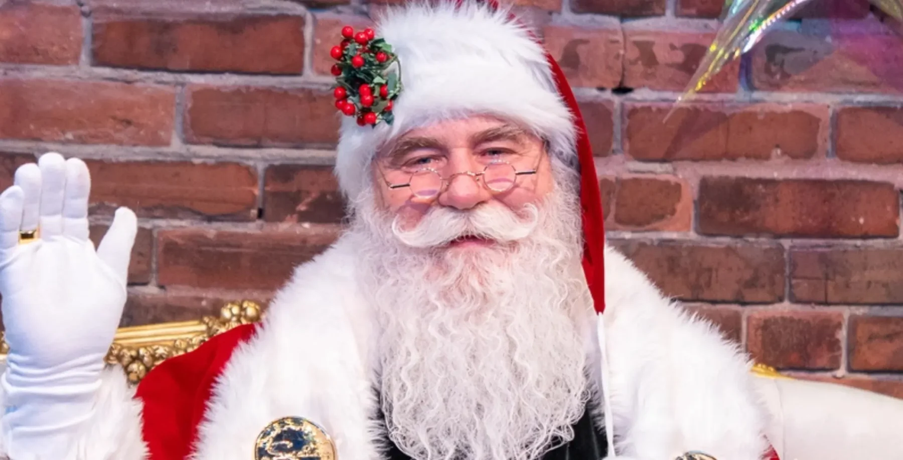 Festive Santa Claus in holiday costume with red and white attire, glasses, and a holly accessory, sitting in front of a brick wall for Christmas celebrations in Toronto.
