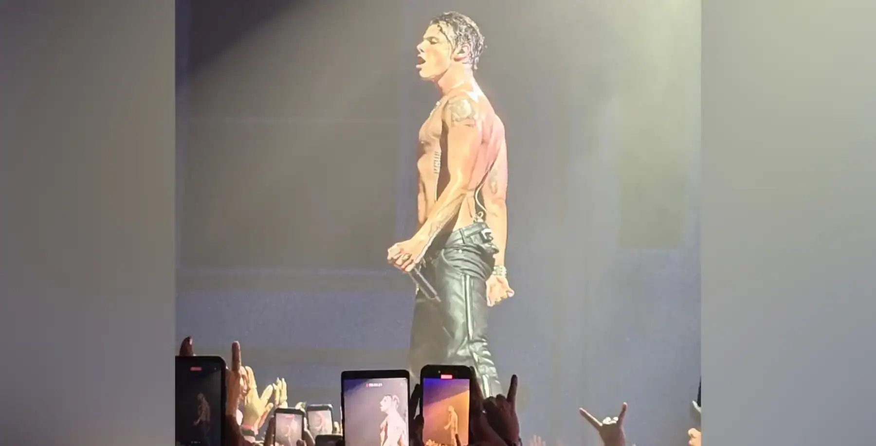 Vibrant concert scene featuring a shirtless male performer with tattoos on stage, captivating the crowd of fans recording with smartphones.