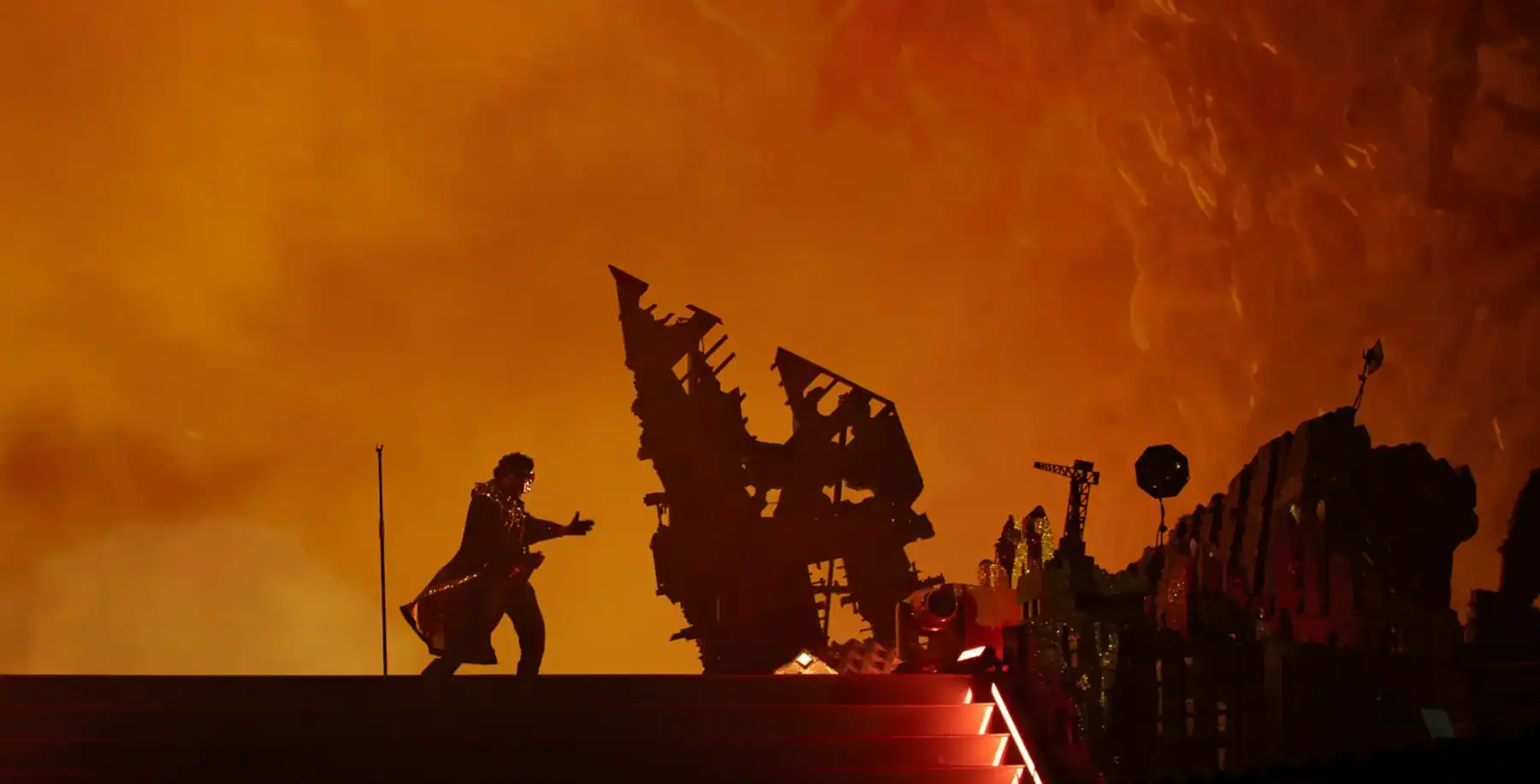 A person performing on stage with a fiery orange background, surrounded by decorative structures and props from NOW Toronto arts and cultural events.
