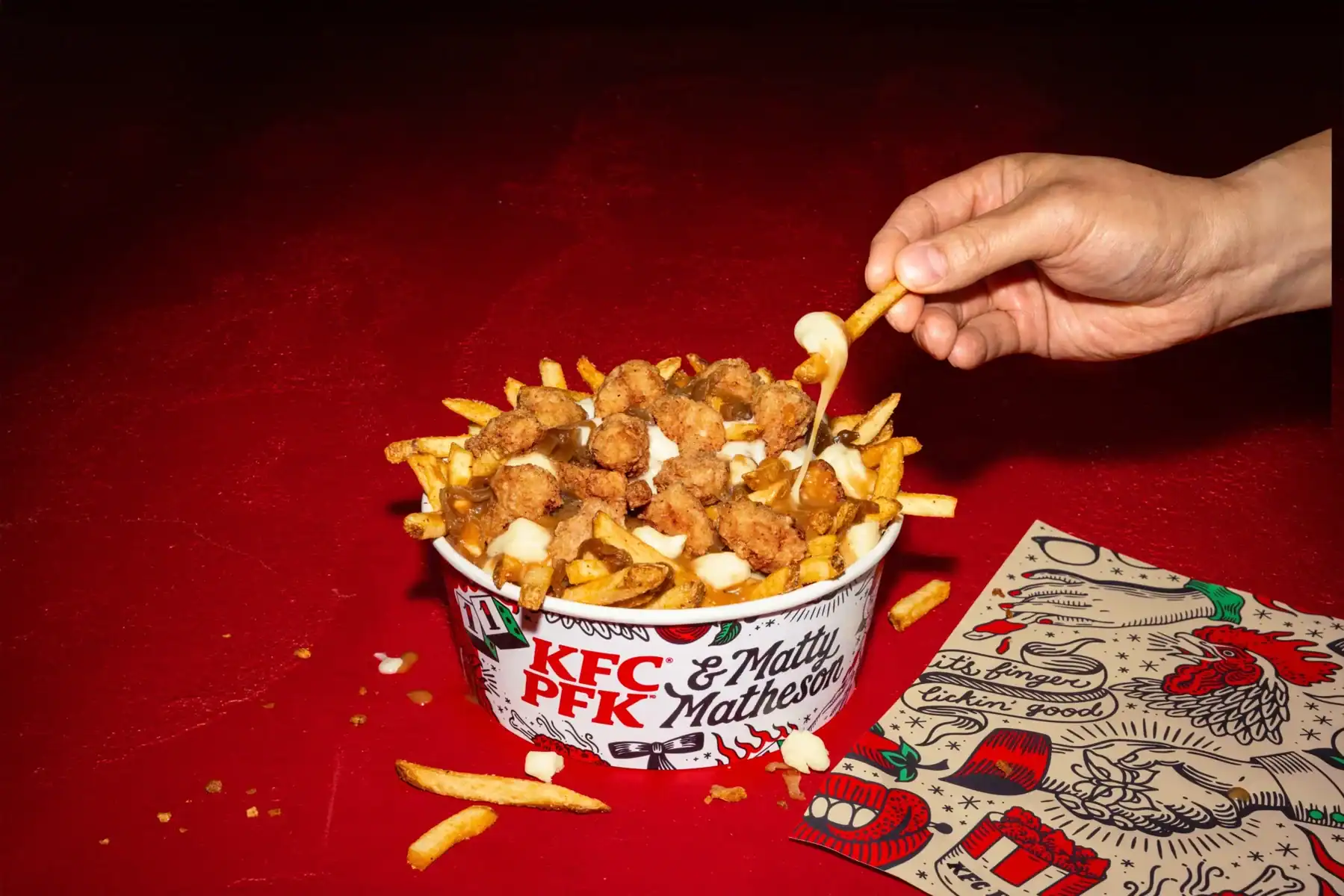 Golden fried chicken bites, fries, and cheese in a KFC and Matty Matheson collaboration bucket on a red surface.