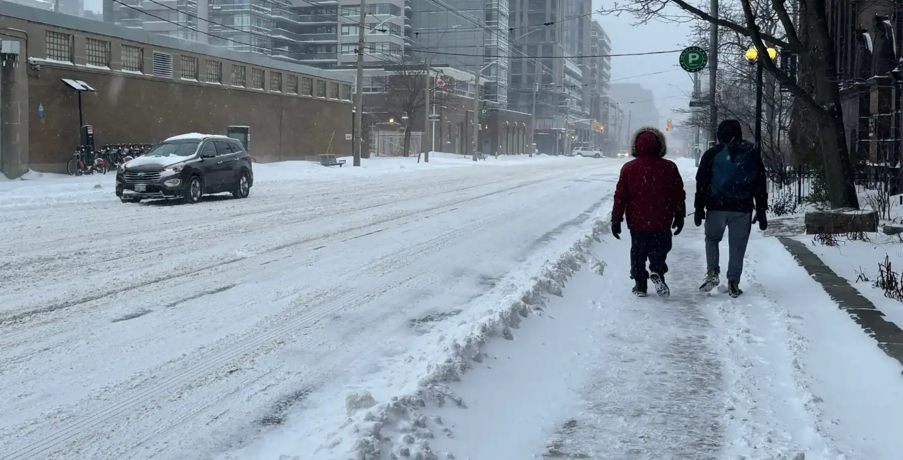 Heavy snowfall in downtown Toronto during winter, with pedestrians walking on snow-covered sidewalks and cars driving through snow-blanketed streets.