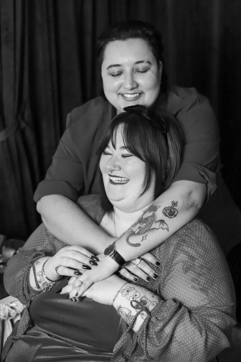 A woman with tattoos hugging another woman with dark hair, both smiling warmly in a black-and-white photo, capturing a moment of friendship or celebration.