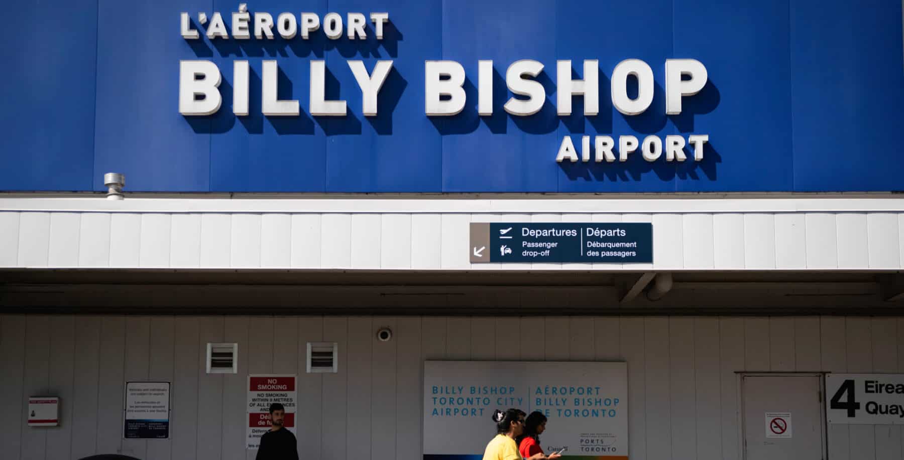 the front of billy bishop airport with a destinations sign and some travellers walking