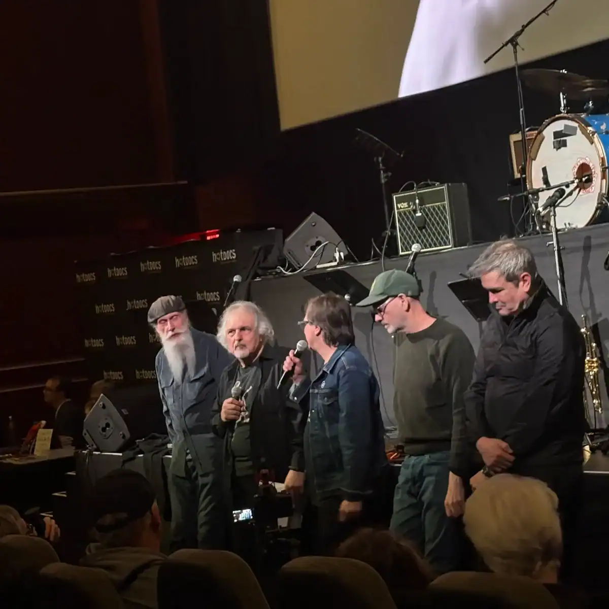 An image of five people on stage at a Toronto event, discussing the life and legacy of Nash The Slash, a pioneering musician from Toronto, with musical instruments and stage equipment in the backgroun.