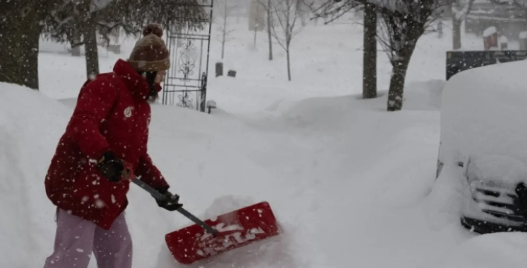 A person in a red winter coat shoveling snow from a sidewalk in Toronto, with snow-covered trees and parked cars in the background.