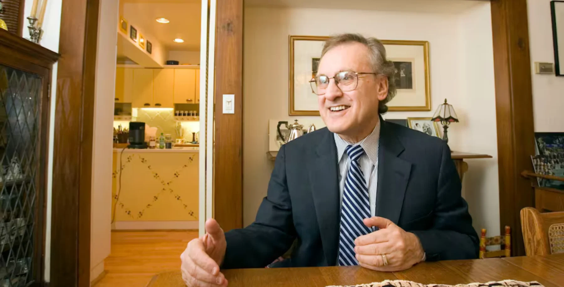 An image of a smiling man in a suit and glasses, sitting at a wooden table in a cozy, well-lit room, reflecting the somber moment of Canadian political leaders mourning the passing of Stephen Lewis.