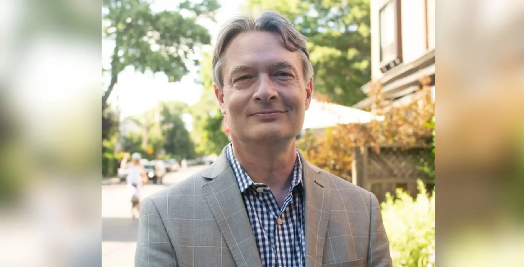 A portrait of a middle-aged man in a gray checkered blazer and blue shirt, standing outdoors with trees and sunlight in the background, representing Toronto's vibrant community and professional enviro.