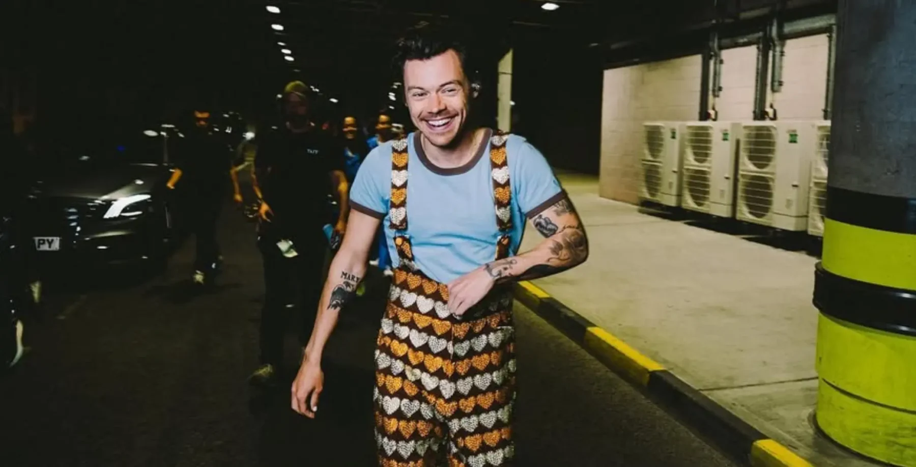 A joyful man with tattoos and a bright smile, wearing a colorful patterned outfit, walking in Toronto during a Pride parade at night, with parade participants and city lights in the background.