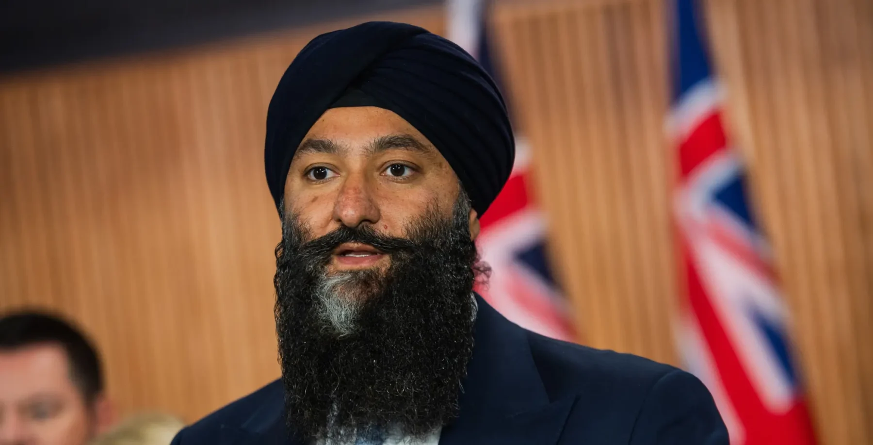 A man wearing a turban and suit speaking at an event, with Canadian flags in the background, during a community gathering related to Toronto's Little Norway Park and Billy Bishop expansion concerns.