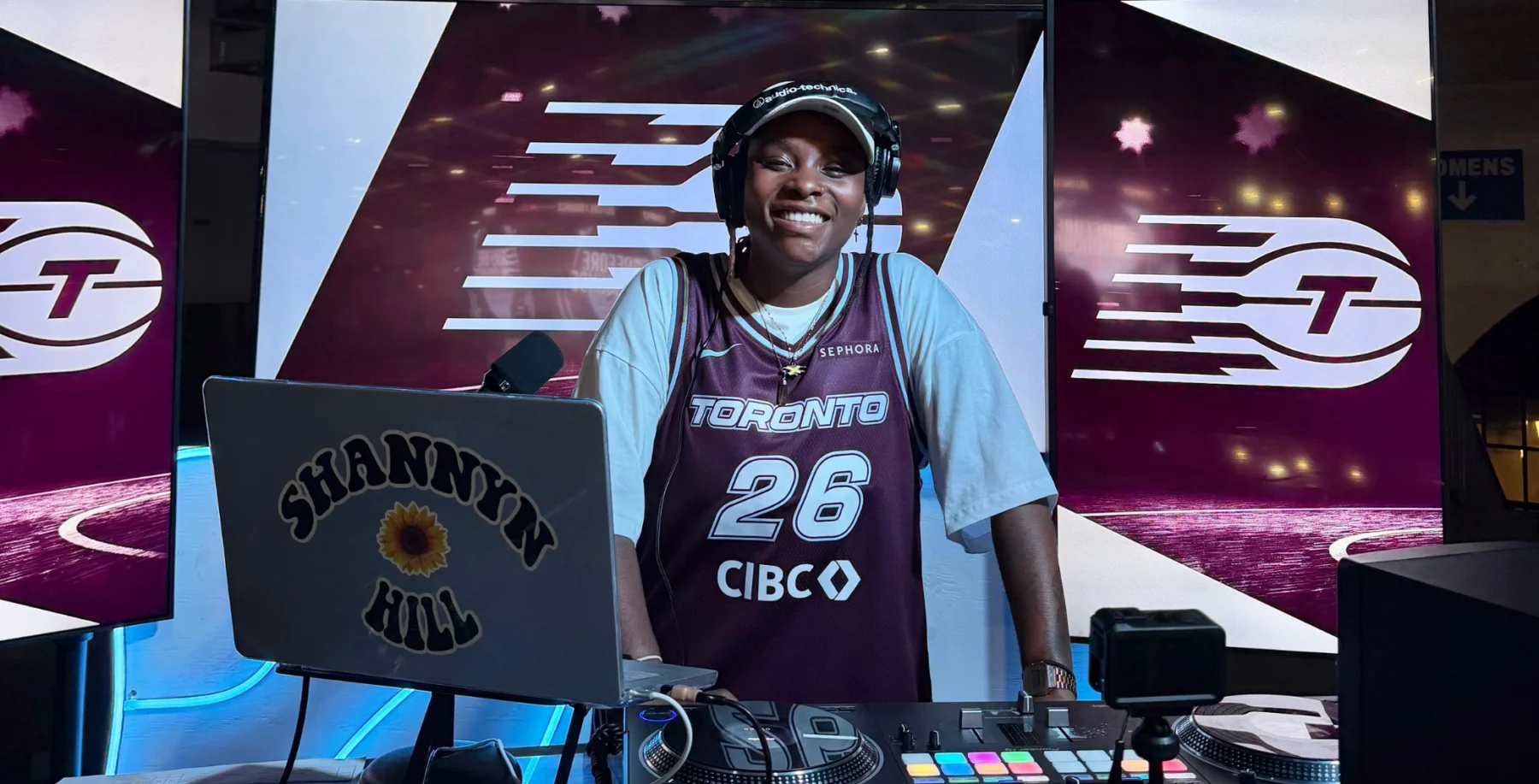 Shannyn Hill, a professional DJ, performing at a Toronto event, wearing a Toronto Raptors jersey, with DJ equipment and vibrant background visuals.