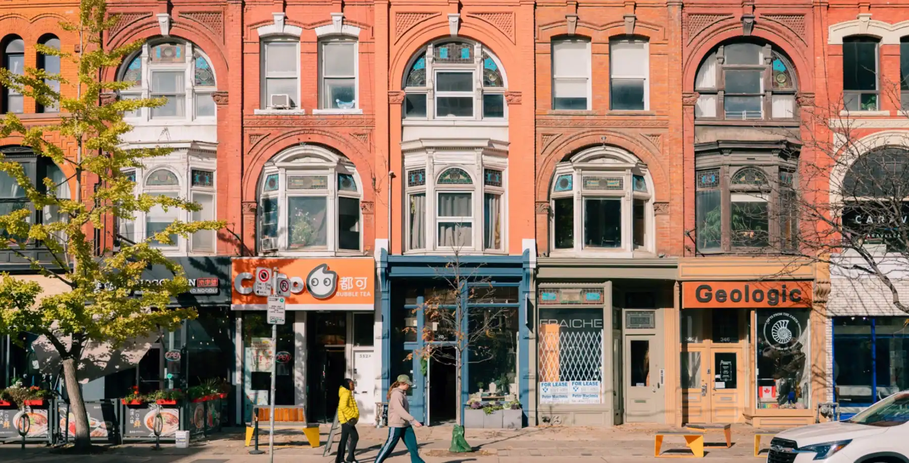 A vibrant Toronto street scene featuring historic architecture, storefronts, and people walking along the sidewalk.