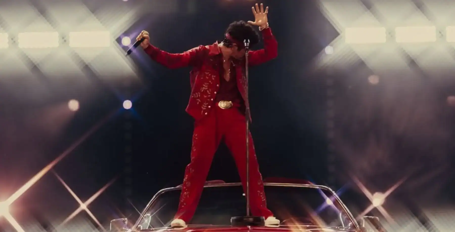 Elvis Presley impersonator in a red jumpsuit performing on stage with a microphone, standing on a car, with bright stage lights and a lively concert atmosphere.