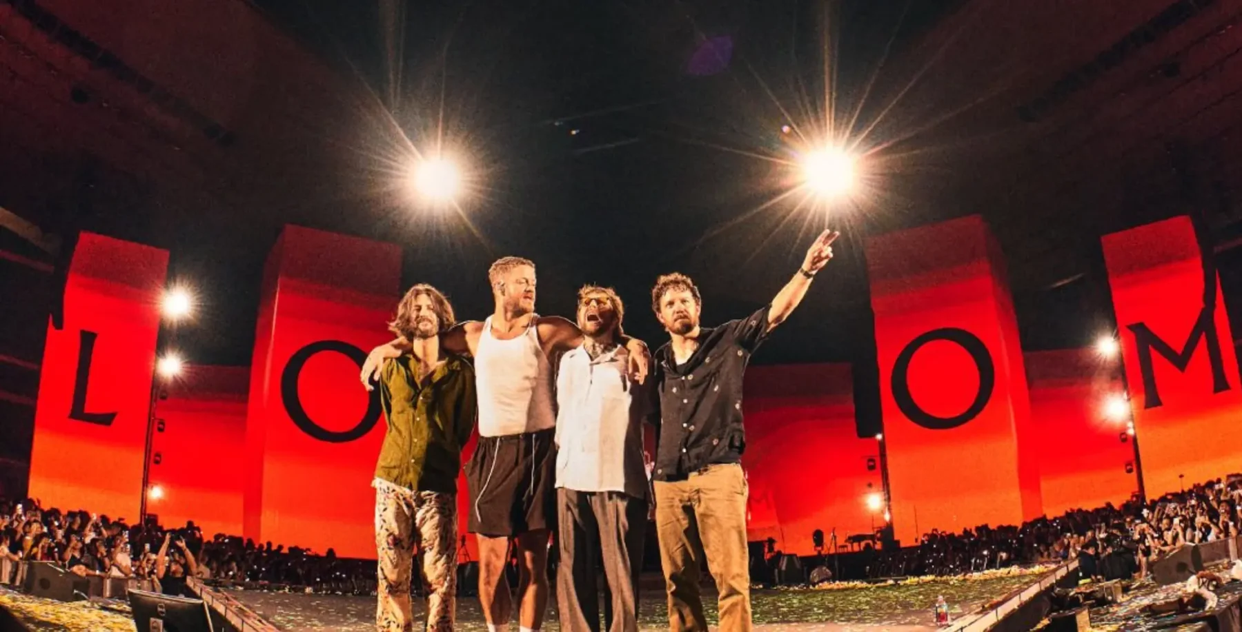 A group of four musicians performing live on stage at the NOW Toronto event, illuminated by bright stage lights, with a large audience in the background.