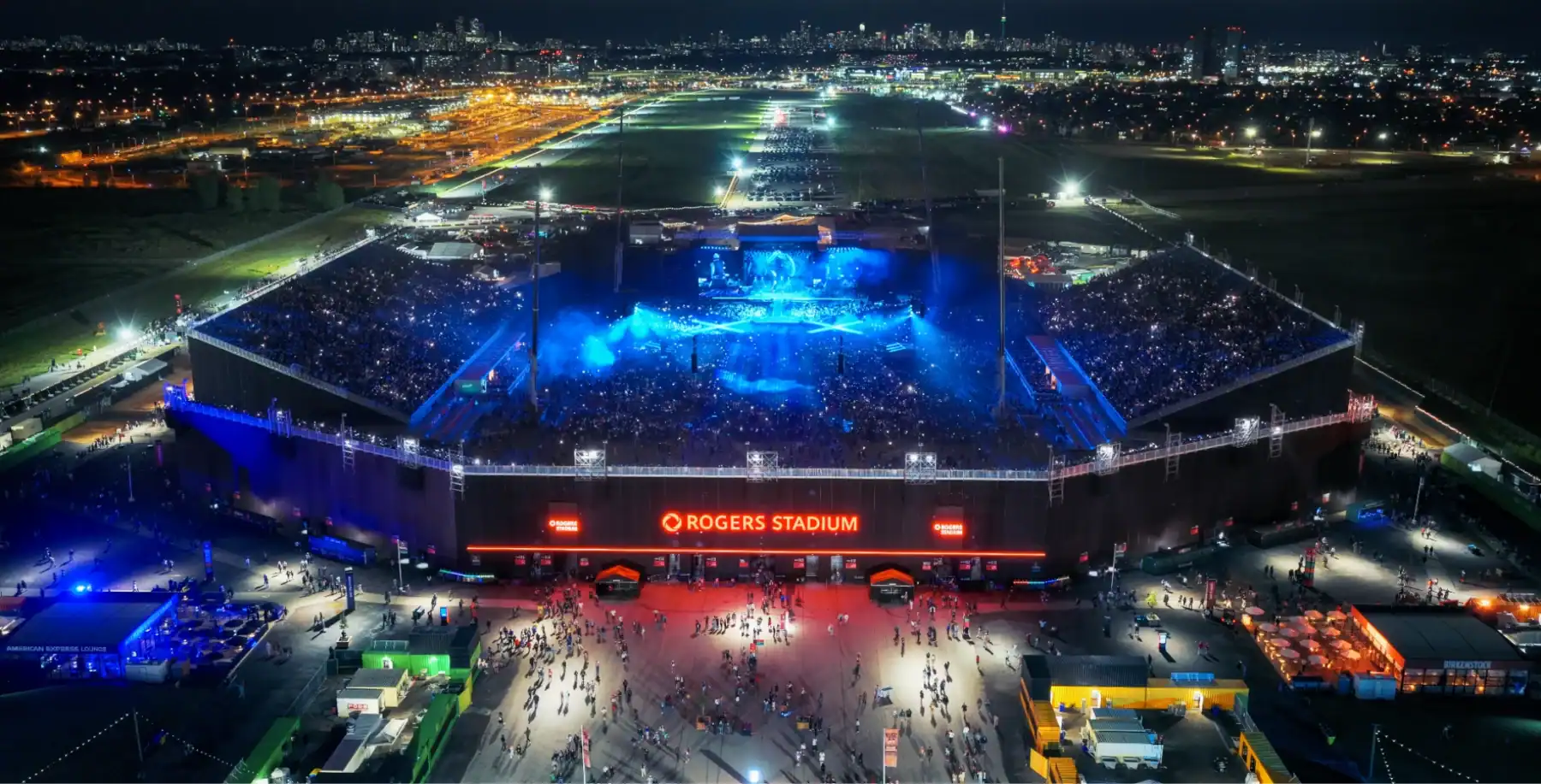 Aerial view of Rogers Stadium illuminated at night during a concert, with a large crowd gathered outside and city lights in the background, showcasing Toronto's vibrant entertainment scene.