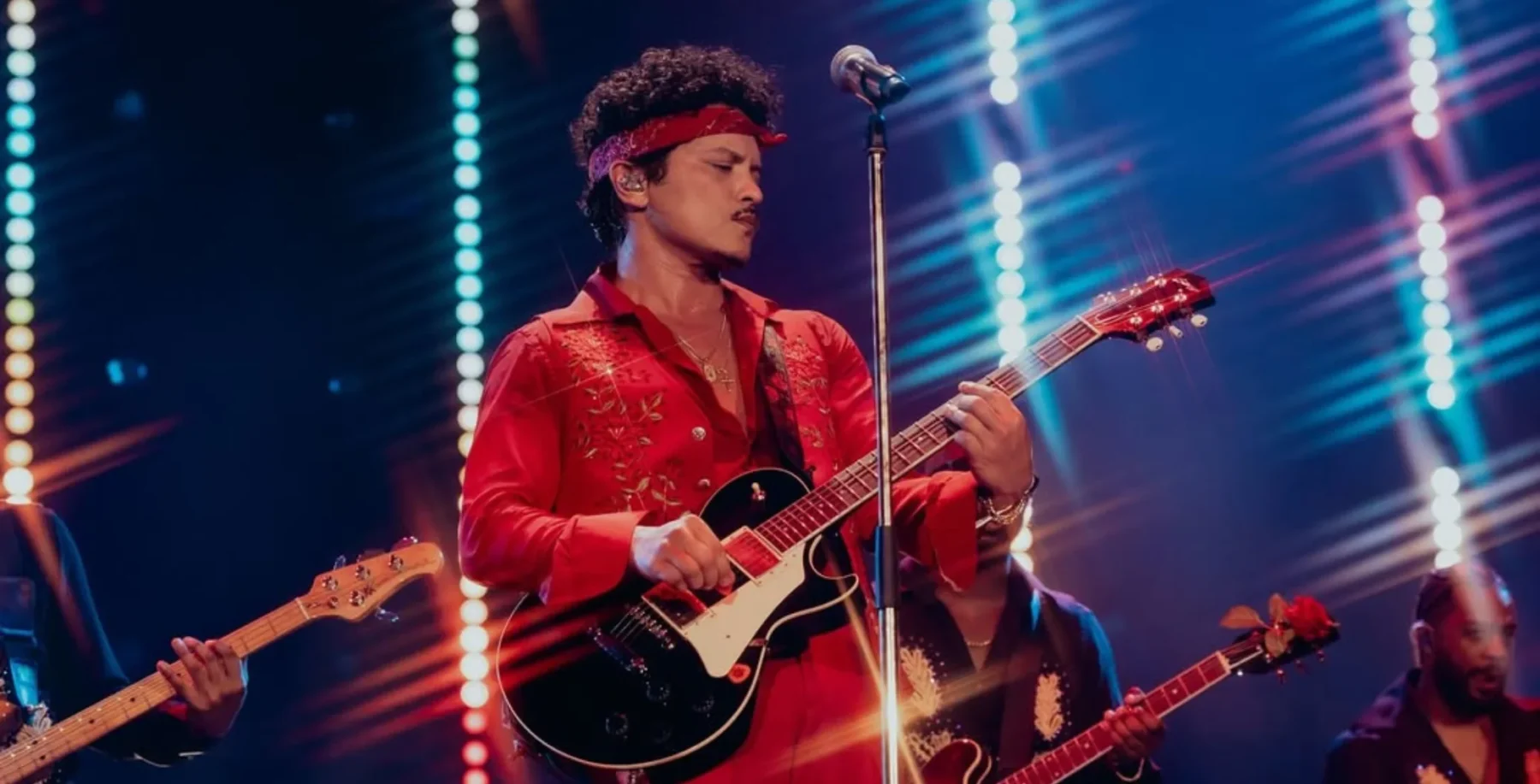 An energetic musician wearing a red shirt and headband plays an acoustic guitar on stage during a live concert at NOW Toronto, illuminated by colorful stage lights.