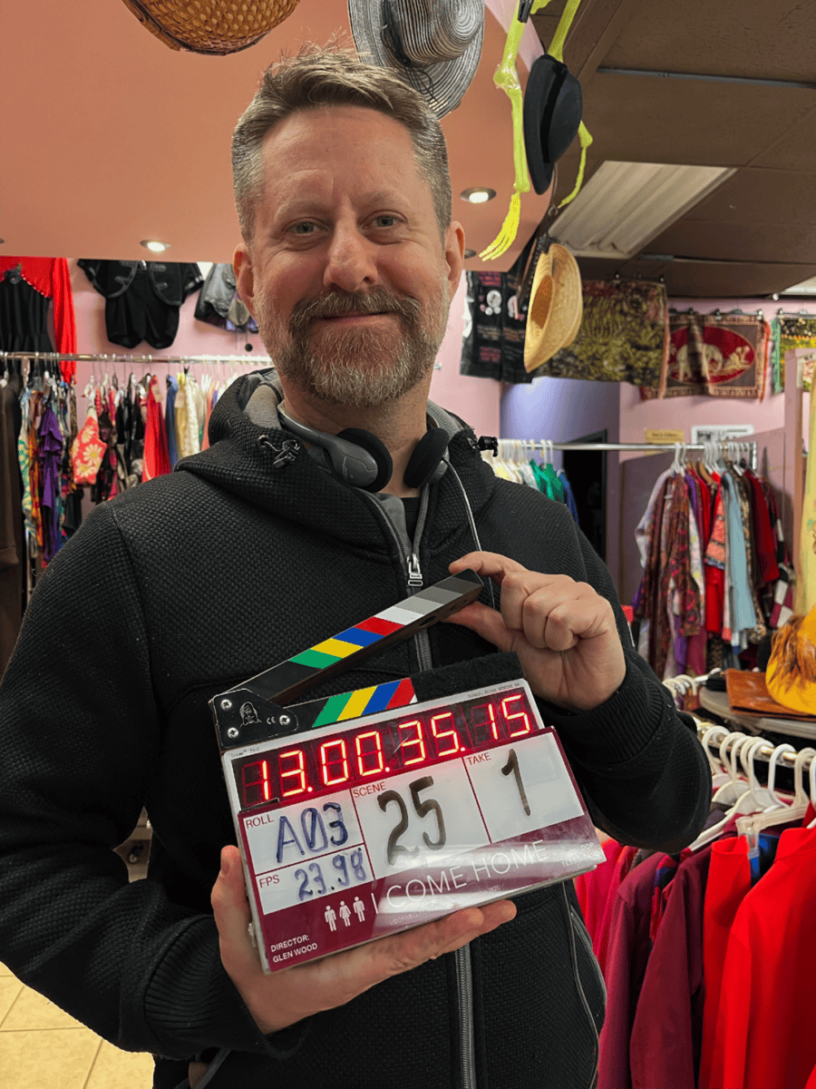 A smiling man with a beard and headphones around his neck holding a clapperboard inside a clothing store, surrounded by colorful apparel and accessories, representing Toronto retail shopping.