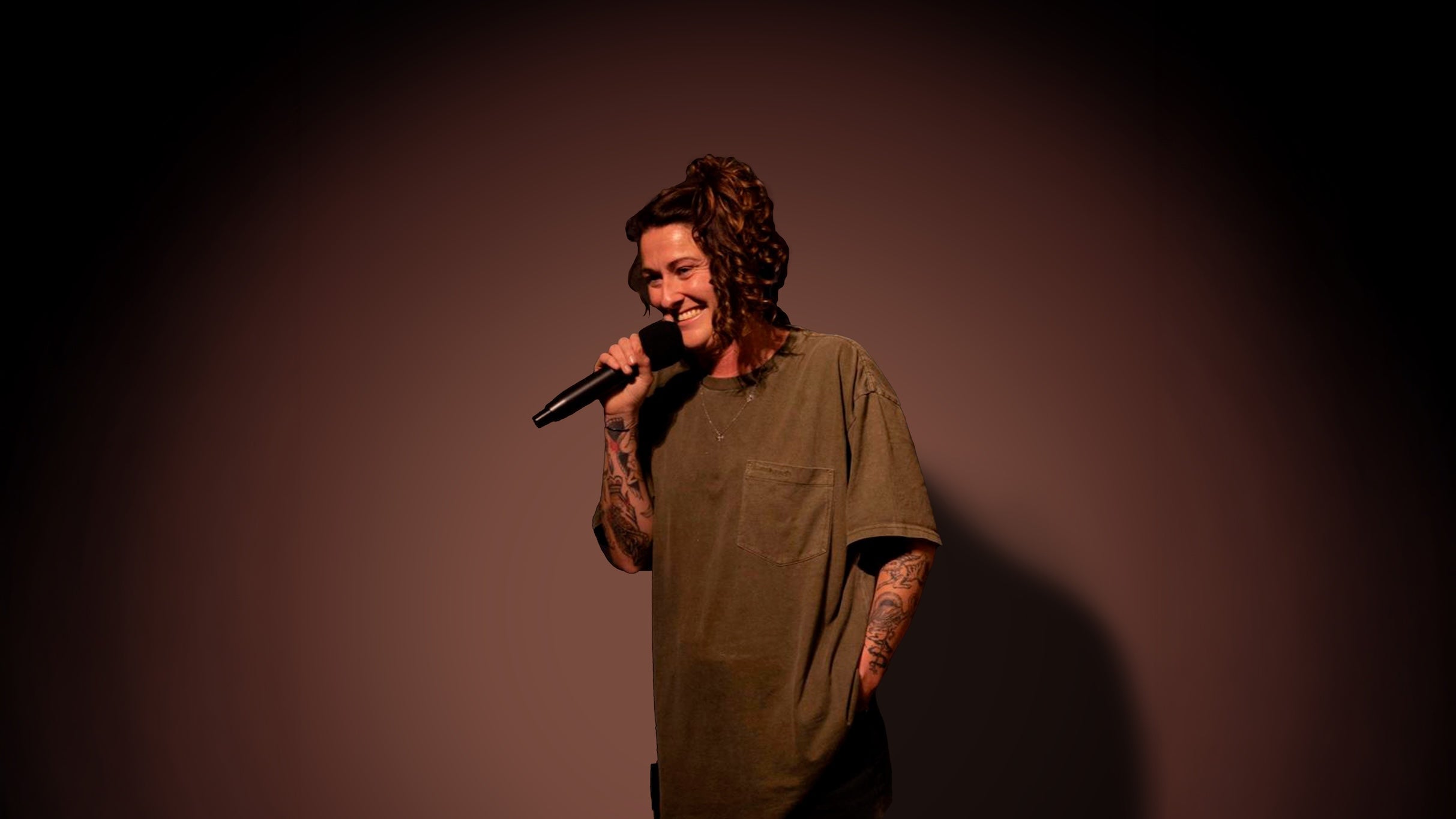 A female comedian with curly hair, wearing an oversized green t-shirt, holding a microphone and smiling while performing on stage at a NOW Toronto event, showcasing entertainment and comedy in Toronto.