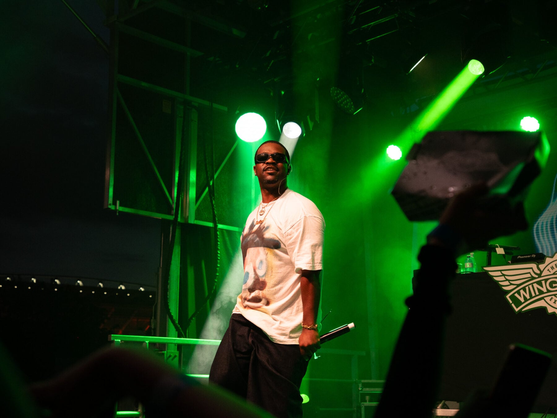 A young performer on stage at a concert in Toronto, illuminated by vibrant green stage lights, holding a microphone, with a DJ setup in the background.