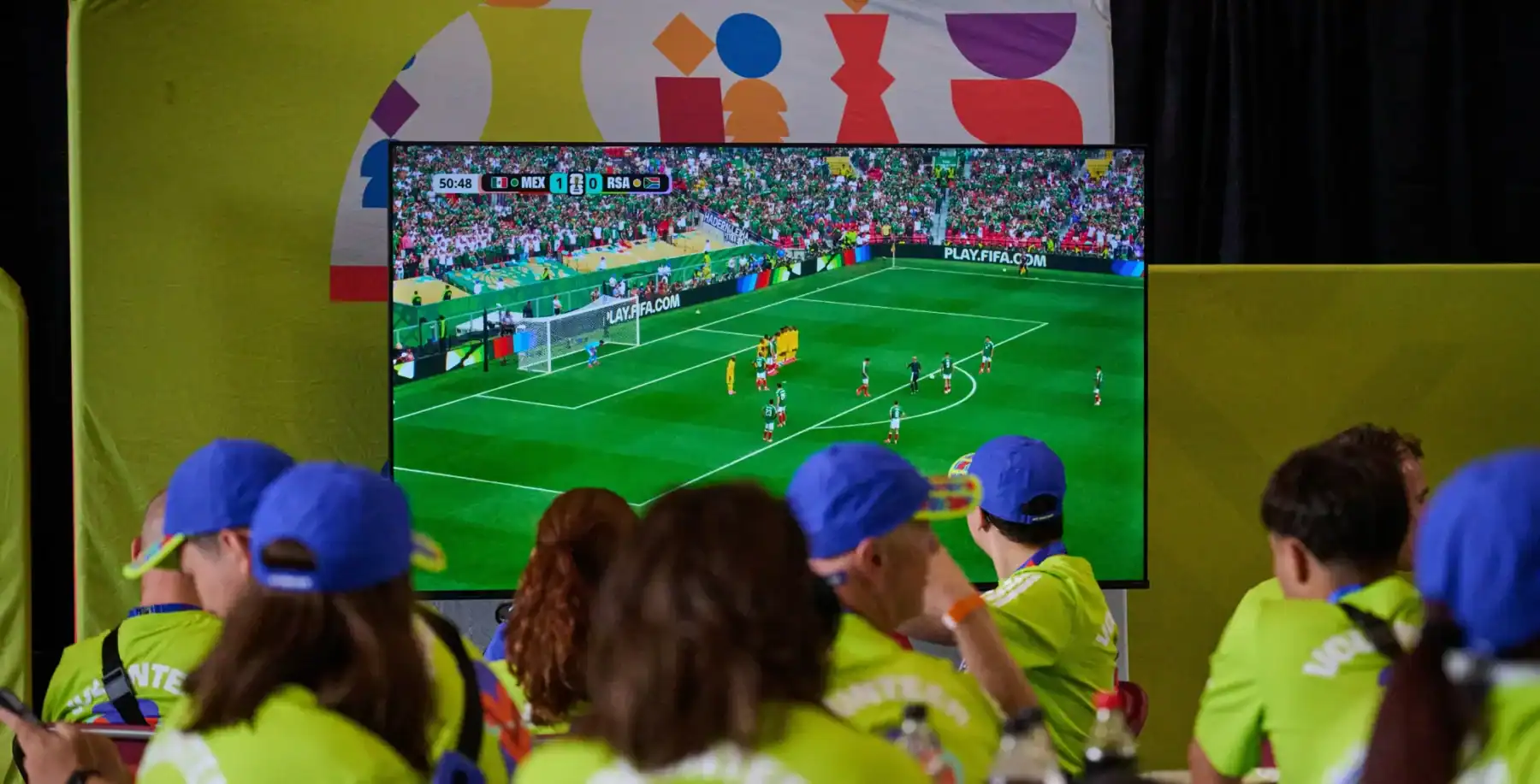 Children watching FIFA World Cup match on a large screen at a watch party in Toronto, wearing blue caps and bright yellow shirts.