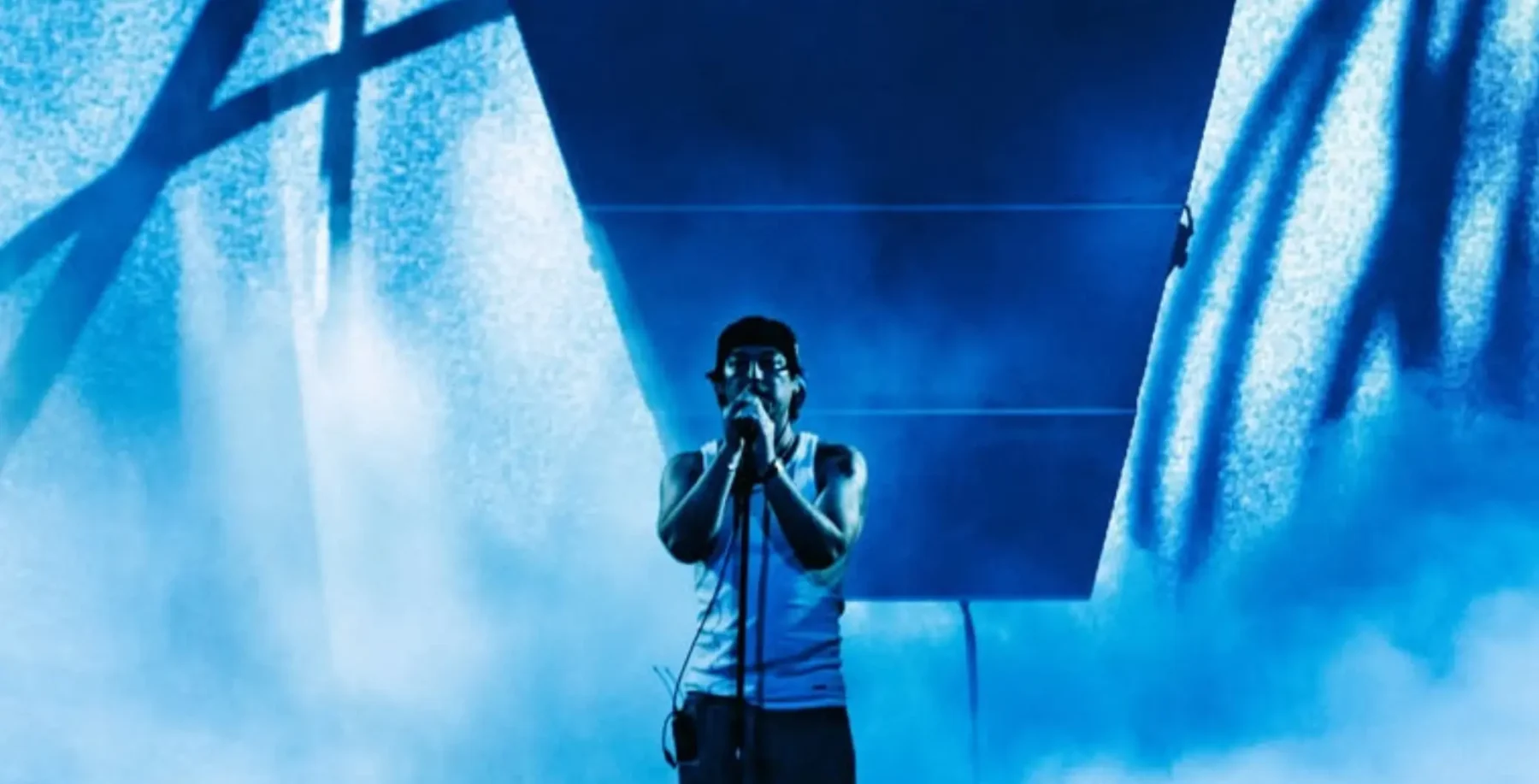 Joji on stage at a Toronto concert, illuminated by blue lights, singing into a microphone with a large screen behind him.