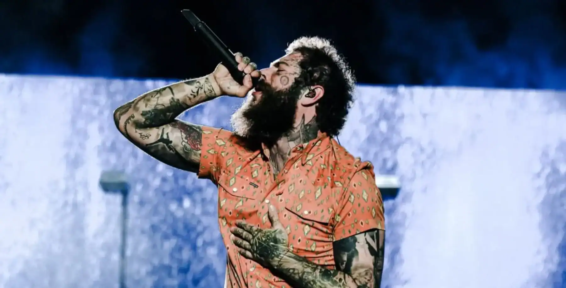 A singer with a full beard and tattoos passionately performing on stage, wearing an orange patterned shirt, under dramatic blue lighting at a live concert in Toronto.