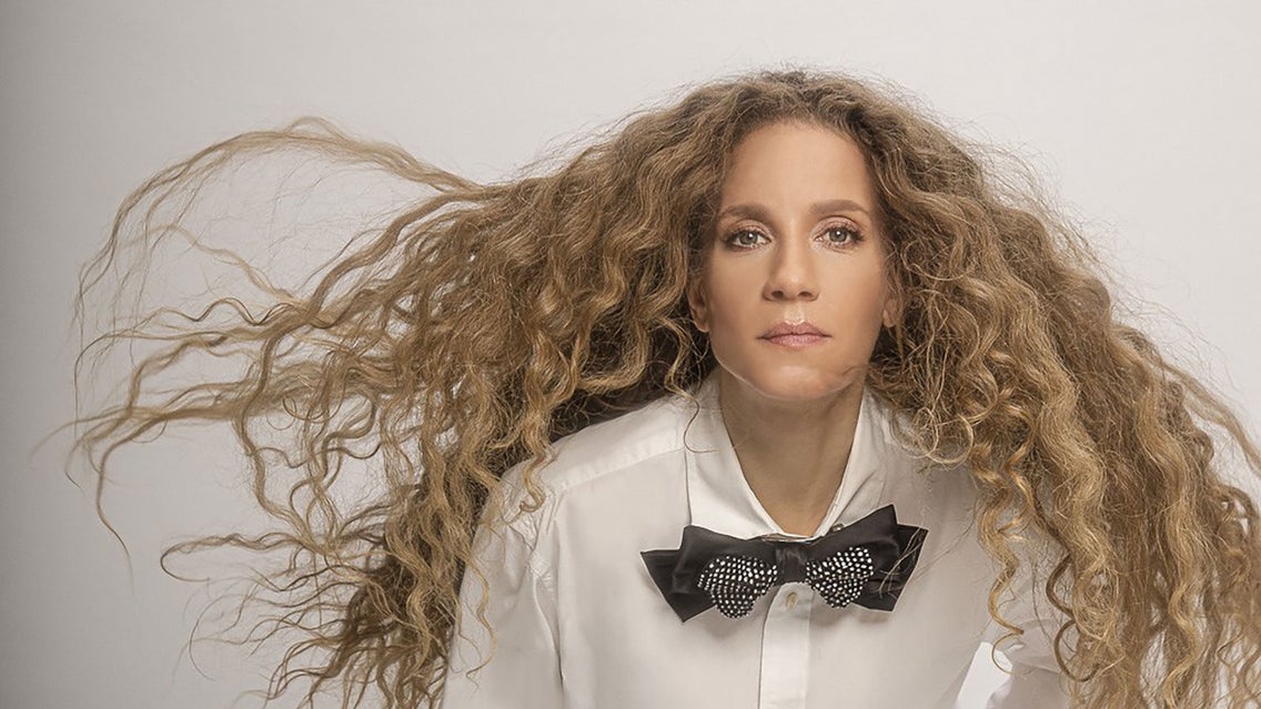 Amanda Marshall with long, curly hair wearing a white shirt and black bow tie, looking directly at the camera for The Dirty 30 Tour.