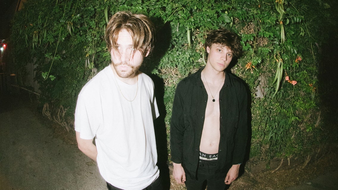 Two young men standing outdoors at night, one in a white t-shirt and the other shirtless with a black jacket, against a lush green background.