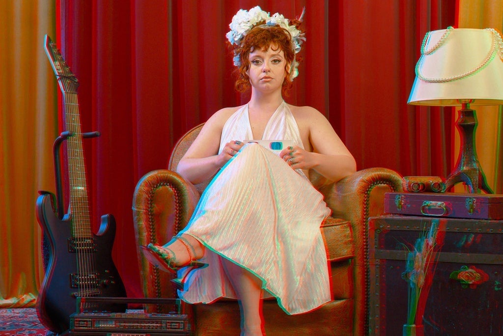 A woman with curly red hair and a floral headpiece sits on a vintage armchair in a colorful room with red curtains, a guitar, and a lamp, portraying a theatrical scene.
